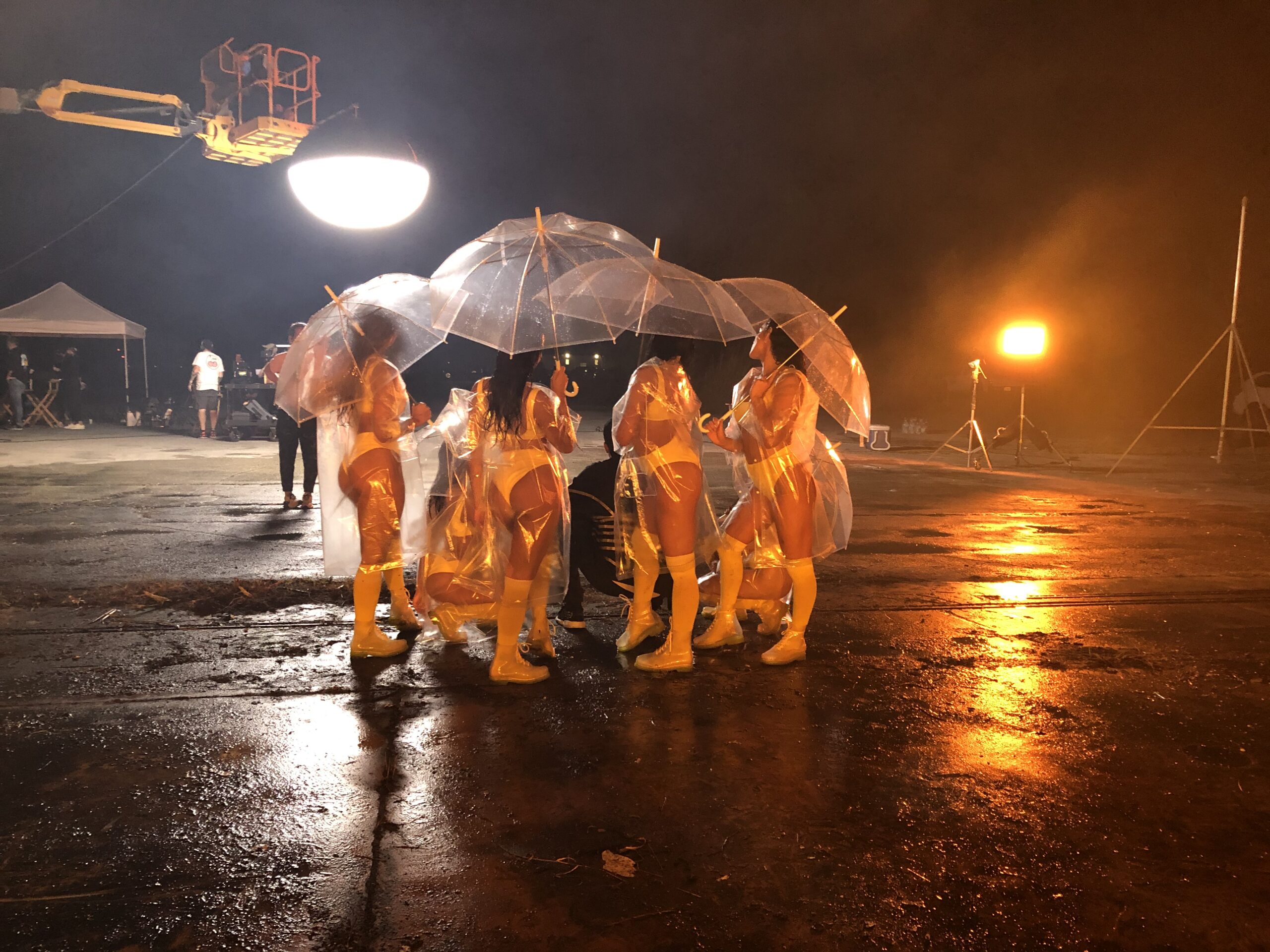 Night scene at an airport featuring several women in white bikinis, boots, raincoats and see through umbrellas standing together in a group. Round overhead Skylight Ballon light and a soft yellow light in the back illuminating the scene.